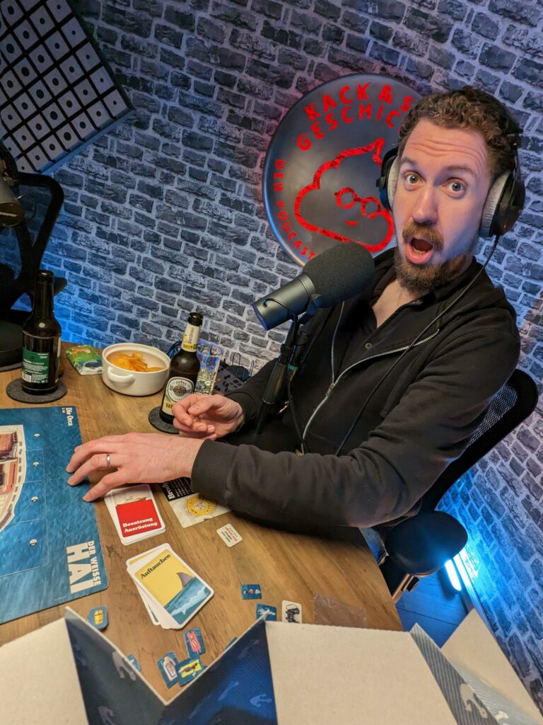 Podcaster Fred sitzt an einem Tisch mit Spielmaterial, Chips und Bier. Im Hintergrund leuchtet das Logo des Podcasts "Kack- und Sachgeschichten".