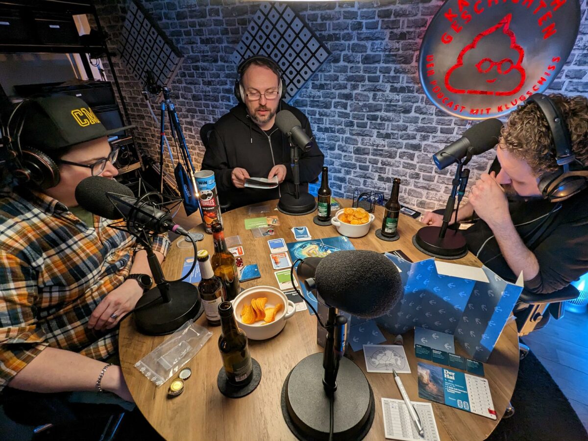 Claudiu, Jörn und Fred (v.l.n.r.) sitzen konzentriert am Spieltisch, während Jörn die Regeln vorliest. Auf dem Tisch stehen mehrere Mikrofone und Bierflaschen, außerdem Chips und das Spielmaterial.