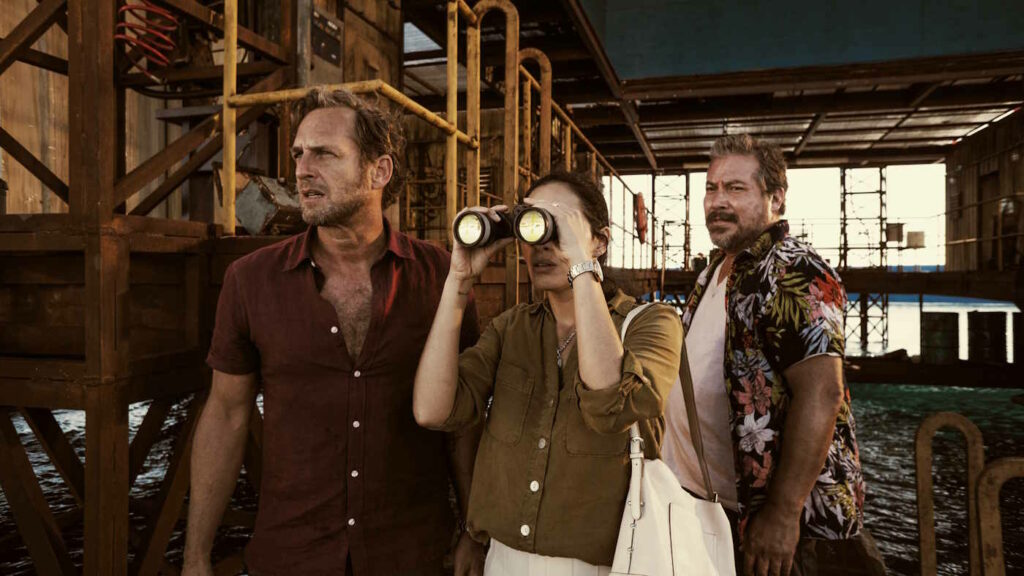 Drei Menschen stehen auf einem Ponton am Anleger einer Ölbohrinsel. Eine von ihnen hält sich ein Fernglas vor die Augen.