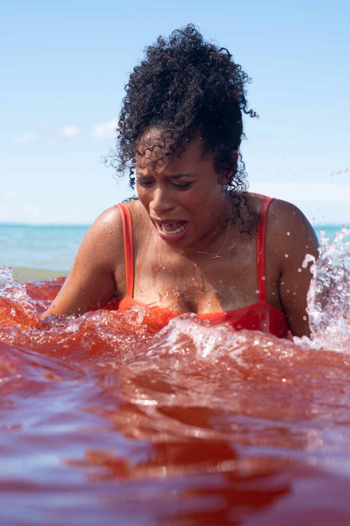 Großaufnahme einer Frau im Badeanzug. Sie steht bis zur Brust im Wasser und schaut verängstigt nach unten. Das Wasser um sie herum ist rot gefärbt.