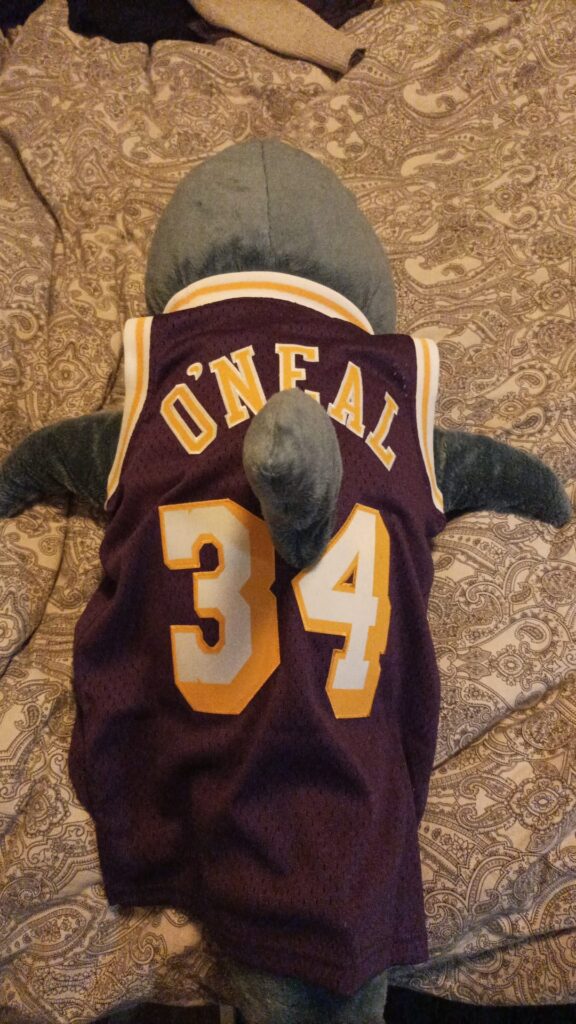 Ein Blahaj liegt auf einem Bett. Er trägt das Basketball-Jersey von Shaquille O'Neal.