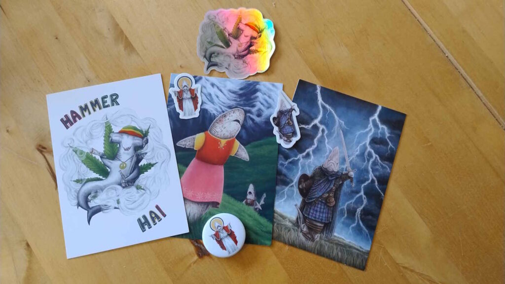 Mehrere Postkarten, Aufkleber und ein Button mit lustigen Hai-Motiven liegen auf einem Tisch.
