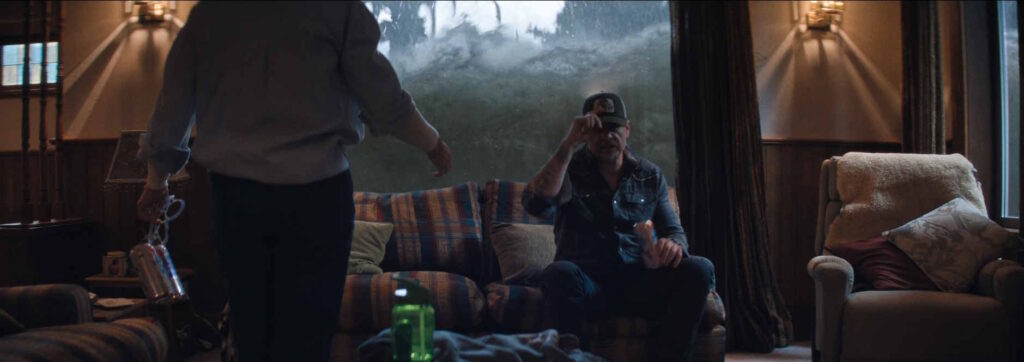 Szenenbild aus dem Film "Thrash". Ein Mann sitzt mit einer Bierdose auf der Couch, vor ihm steht eine Person mit dem Rücken zur Kamera. Am Wohnzimmerfenster hinter ihm steht das Wasser mannshoch.